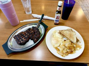 Grilled steak on a sizzling platter, accompanied by eggs, potato hash, and toasted bread. A bottle of A1 sauce and drinks included.
