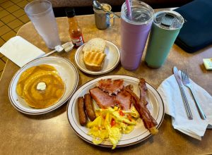 A hearty breakfast plate featuring scrambled eggs, bacon, sausage, and ham, alongside buttered pancakes and toast, with beverages and condiments on the table.