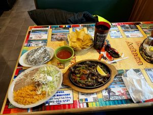 A vibrant Mexican meal featuring sizzling fajitas with peppers, rice, and beans, accompanied by chips and salsa, on a colorful table.